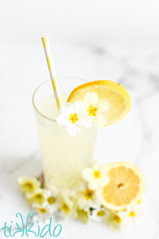 Glass of freshly squeezed lemonade, with a yellow and white straw, and garnished with a slice of lemon and yellow and white edible flowers.