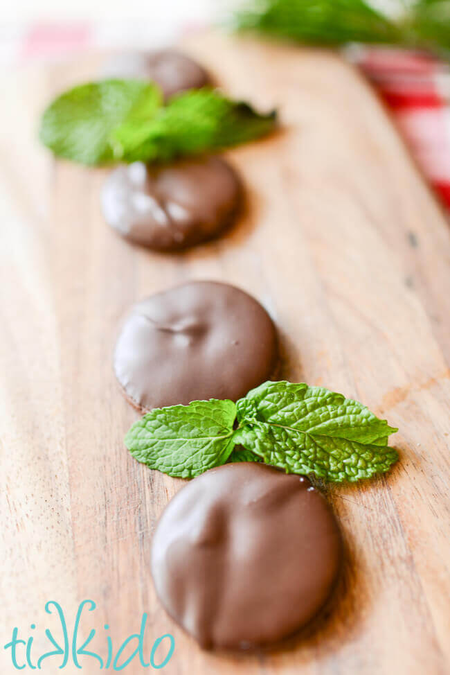 Homemade thin mints in a row on a wooden cutting board, with fresh mint leaves.