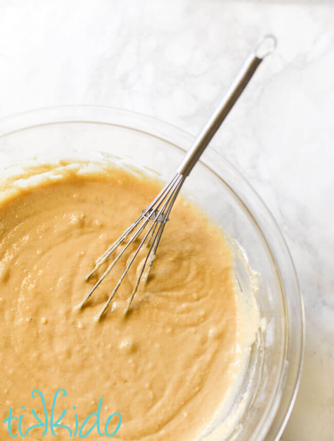 Honey chamomile cake batter in a clear pyrex bowl with a metal whisk, on a white marble background