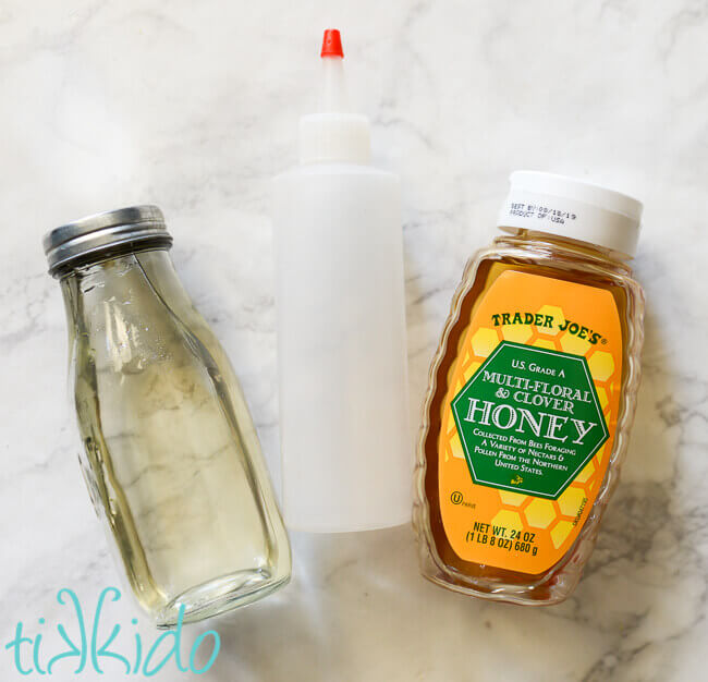 Bottle of simple syrup, empty squeeze bottle, and bottle of honey on a white marble backdrop