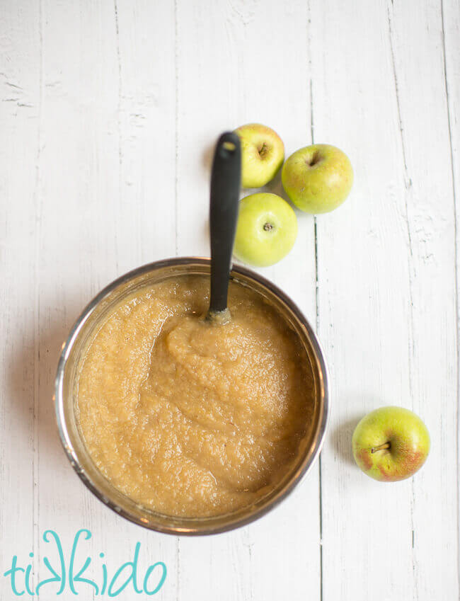 Silver bowl of homemade applesauce on a white wooden surface, with four green apples on the table by the bowl.