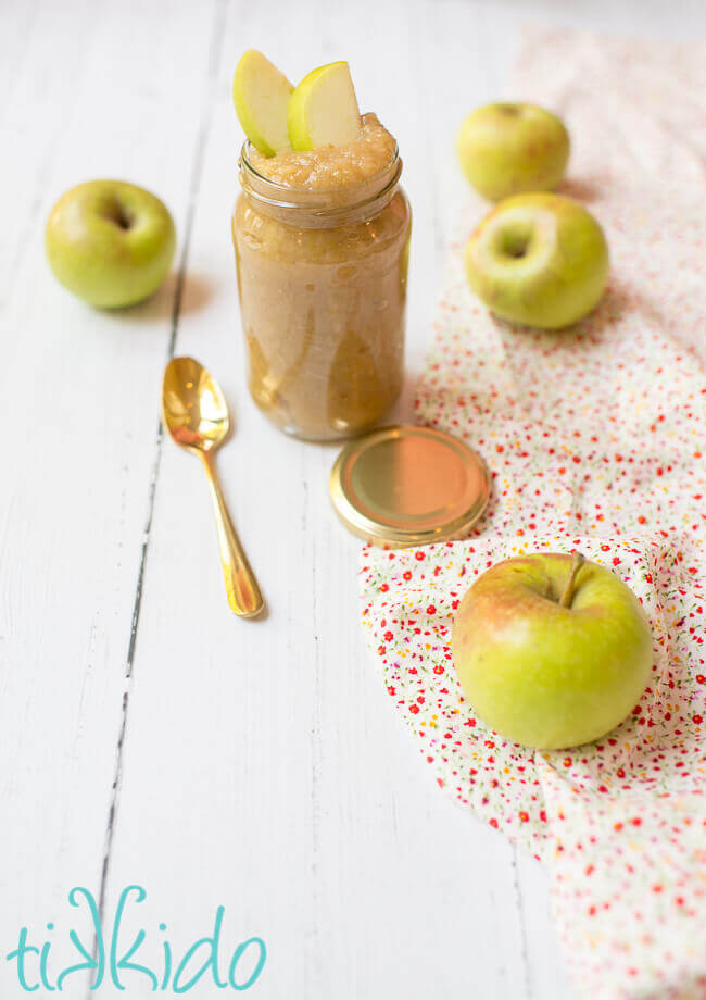Instant pot applesauce in a clear jar, surrounded by fresh apples on a white wooden table.