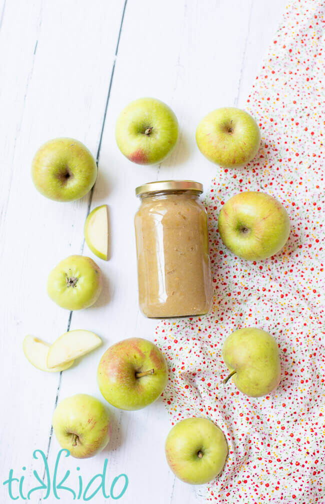 Instant pot applesauce in a clear jar, surrounded by fresh apples on a white wooden table.