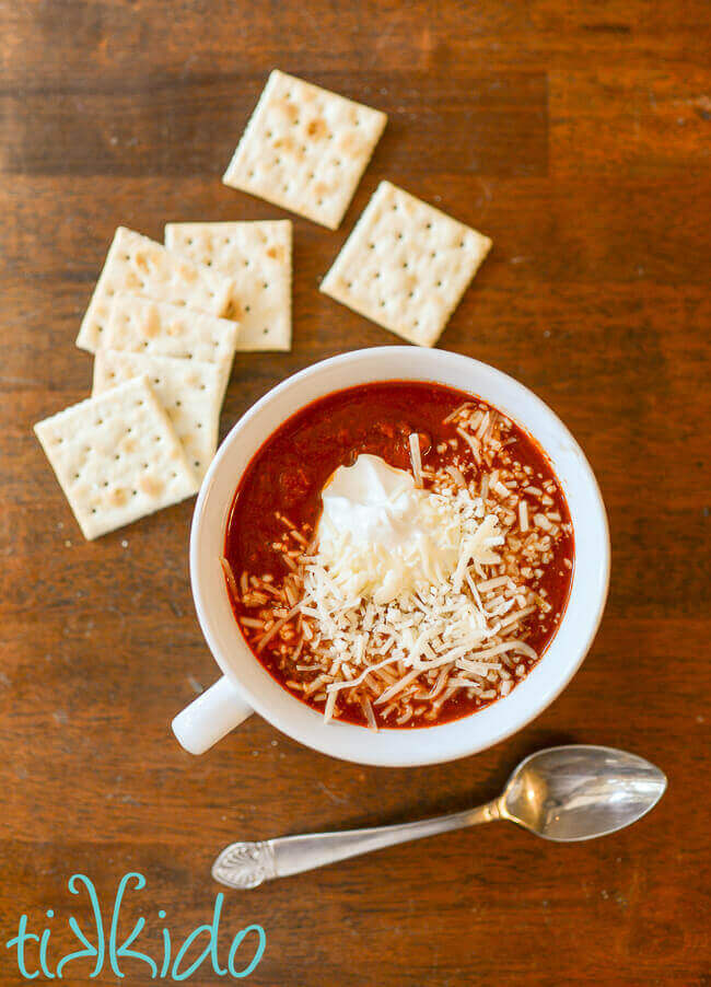 No bean Instant Pot chili in a white bowl topped with sour cream and shredded cheese, surrounded by saltine crackers and a spoon.