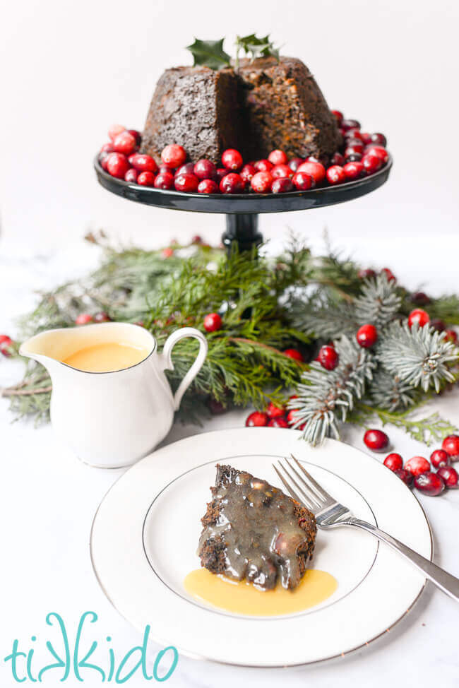 Slice of steamed Christmas pudding covered in hard sauce, in front of a Cranberry Christmas pudding on a cake stand.