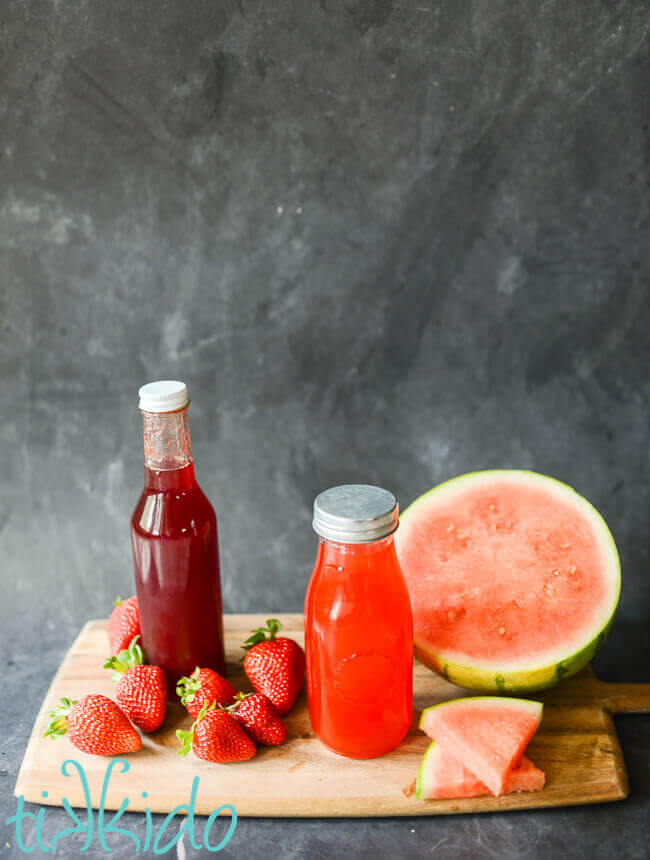 Bottle of watermelon syrup and bottle of strawberry syrup on a wooden cutting board surrounded by fresh strawberries and watermelon.