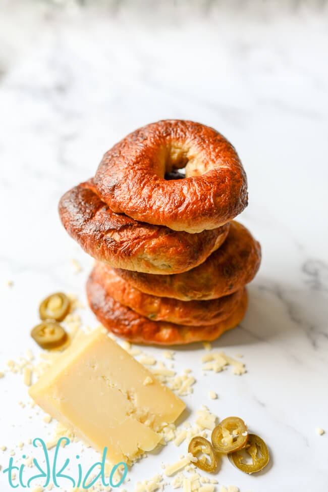 Stack of Jalapeño Cheddar Bagels next to a wedge of cheddar cheese and slices of Jalapeño peppers.