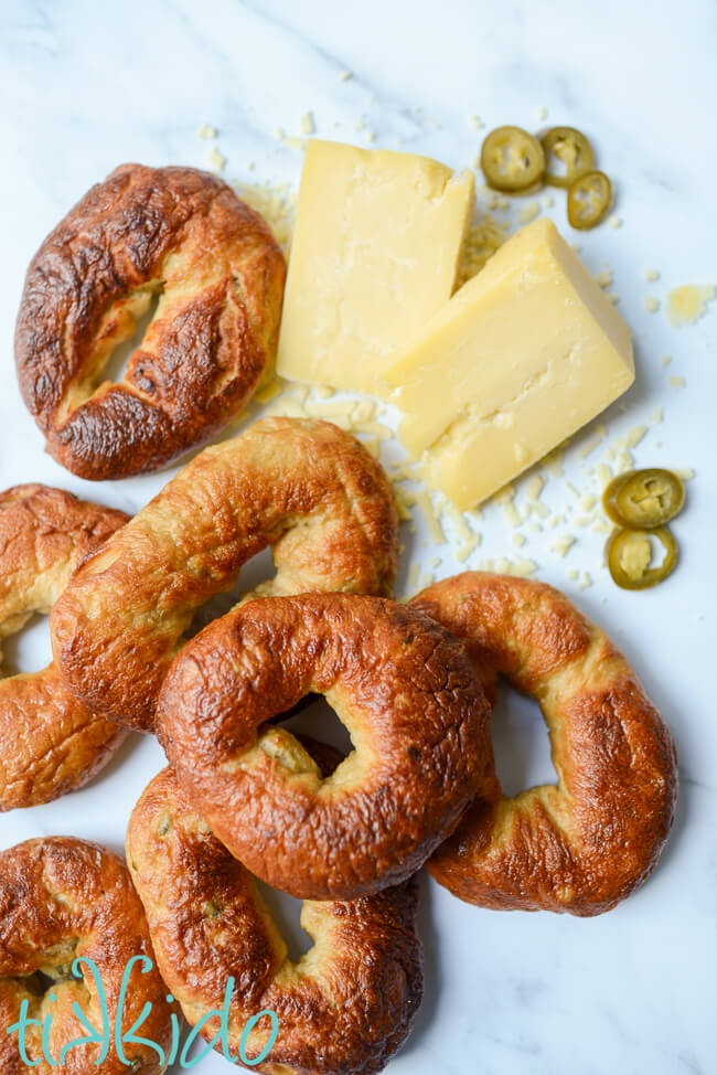 Pile of Jalapeño Cheddar Bagels on a marble surface next to two wedges of cheddar cheese and slices of jalapeno peppers.