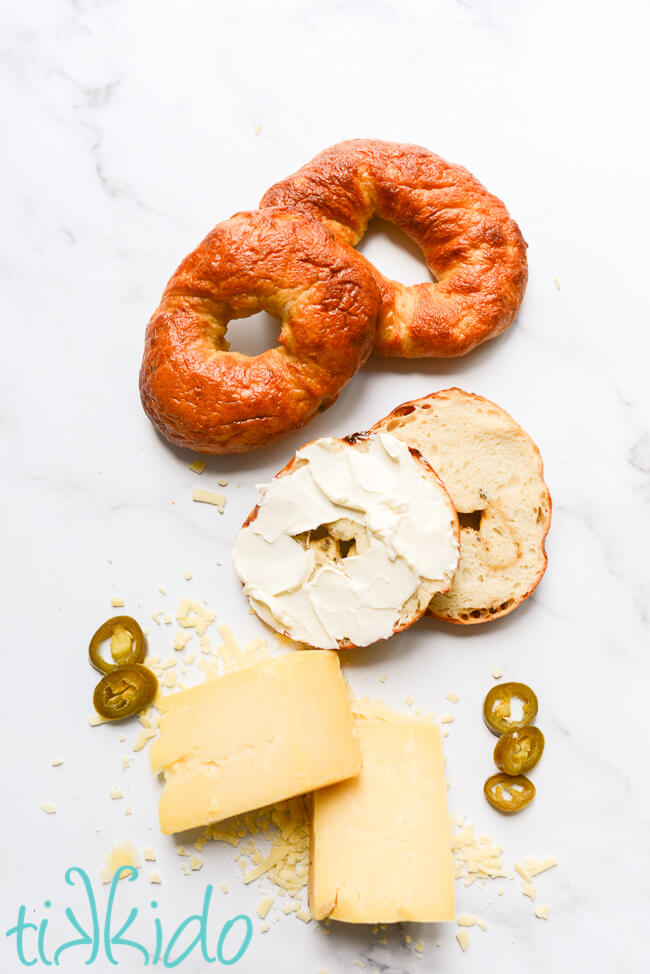 Three Jalapeño Cheddar Bagels, one sliced and spread with cream cheese, next to cheddar cheese and Jalapeño slices.