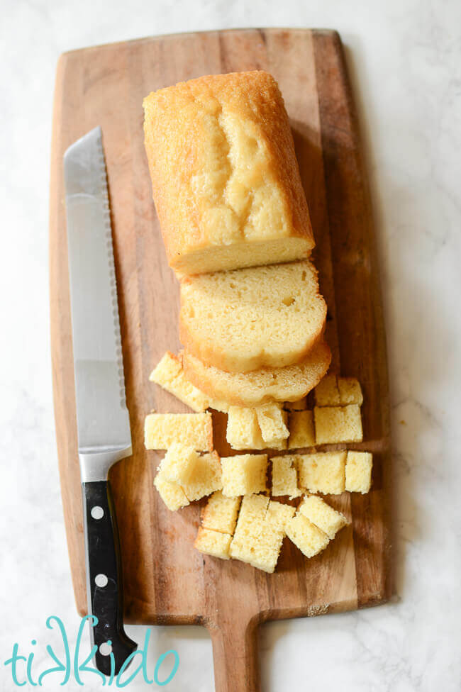 Pound cake, partially sliced, partially cut into cubes, bread knife beside cake, on a wooden cutting board.