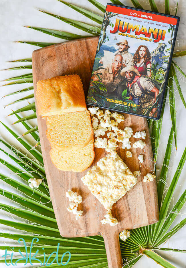 Jumanji DVD, Pound cake, popcorn, and pound cake popcorn marshmallow bars on a wooden cutting board on top of palm fronds.