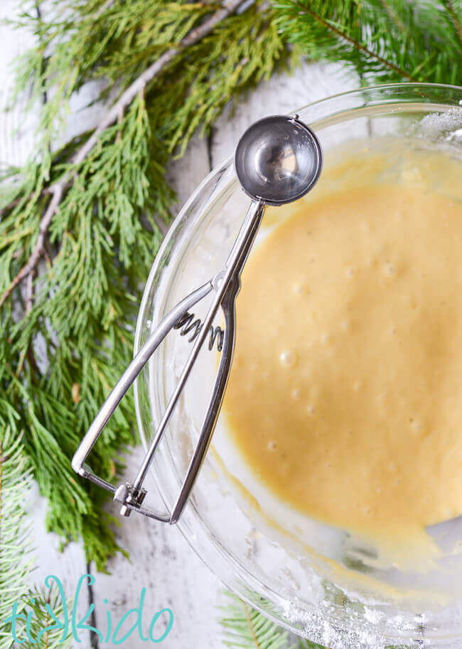 Krumkake batter in a glass bow with a cookie scoop balanced on the rim of the bowl.