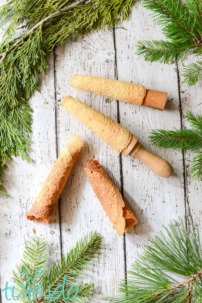 Two rolled krumkake and two krumkake cookies rolled around wooden forms on a white weathered wood background.