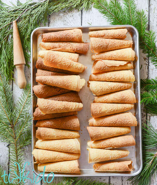 Tray of golden brown Krumkake surrounded by fresh evergreen branches and a wooden krumkake rolling cone form.