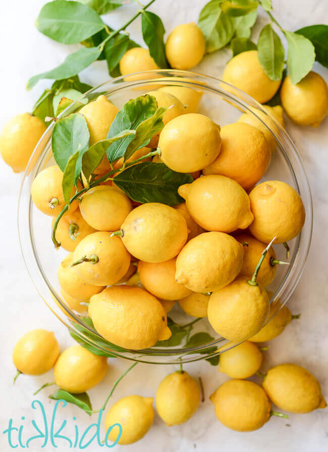 Fresh lemons in a clear glass bowl, surrounded by more lemons on a white marble surface.