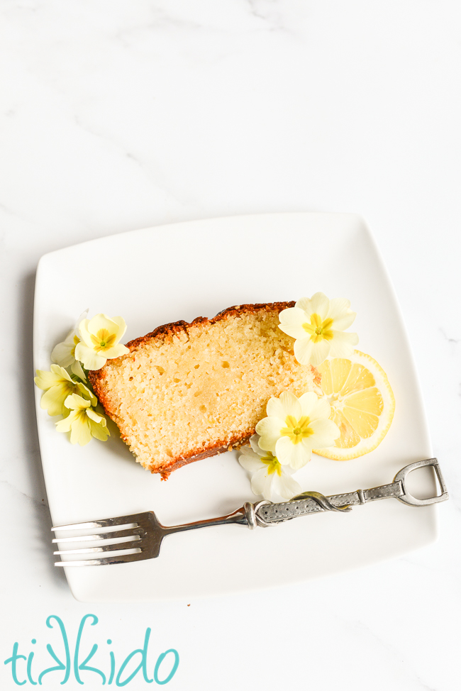Slice of lemon loaf cake garnished with yellow and white edible flowers and a slice of lemon, on a white plate next to a dessert fork.