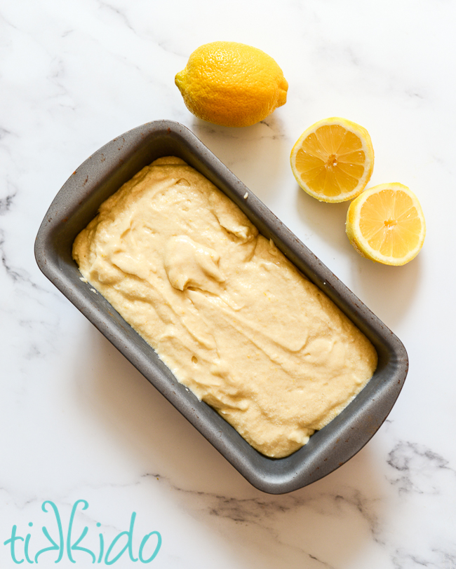 Lemon loaf cake batter spread in a bread pan, ready to be baked, sitting on a white marble surface beside one whole lemon and one lemon cut in half.