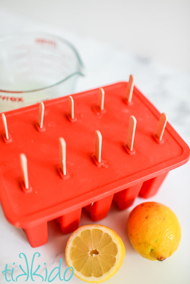 Lemonade popsicles in a red silicone popsicle mold, next to a pyrex measuring cup and two lemons.