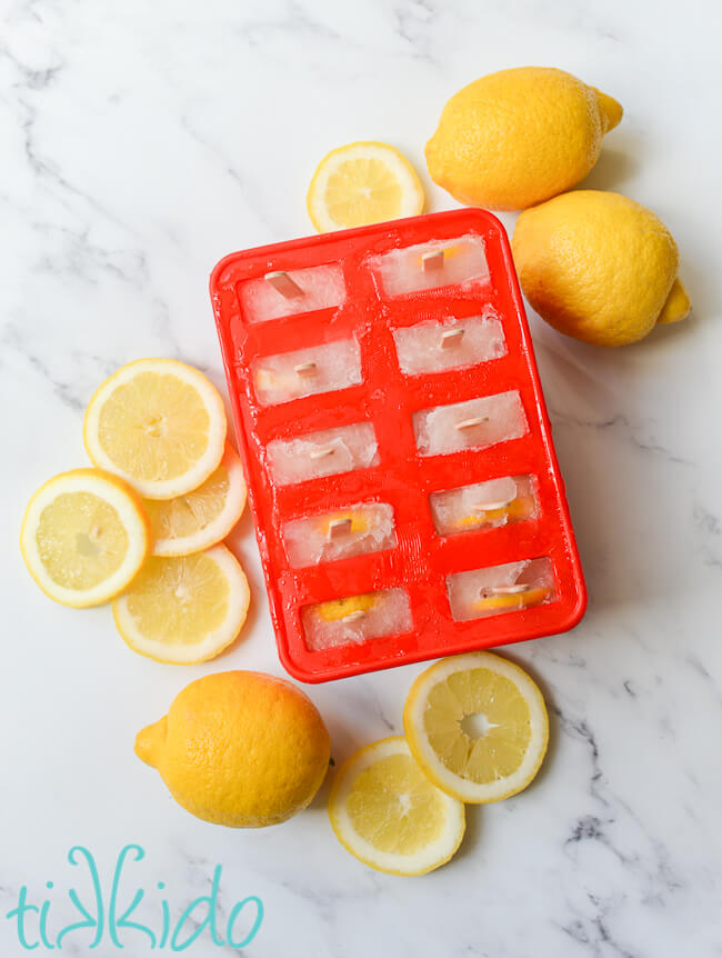 Red silicone popsicle mold filled with lemonade popsicles, sitting on a white marble surface, surrounded by lemon slices and whole lemons.