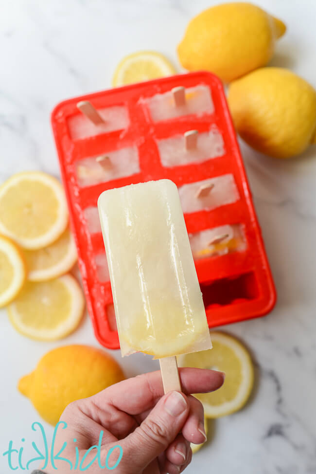 Hand holding a lemonade popsicle above a red popsicle mold filled with lemon popsicles.