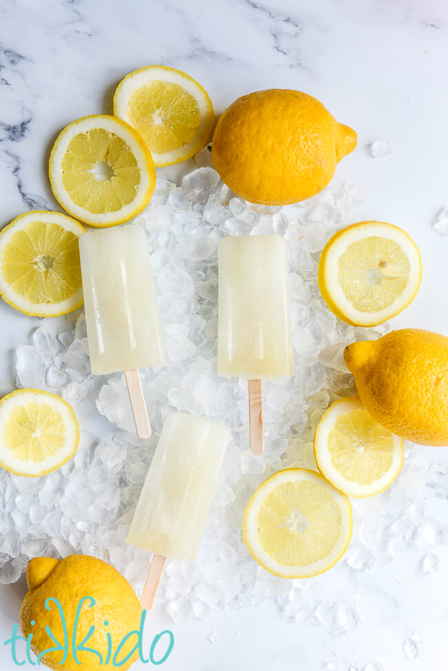 Three lemon popsicles on a bed of crushed ice, surrounded by lemon slices and whole lemons.