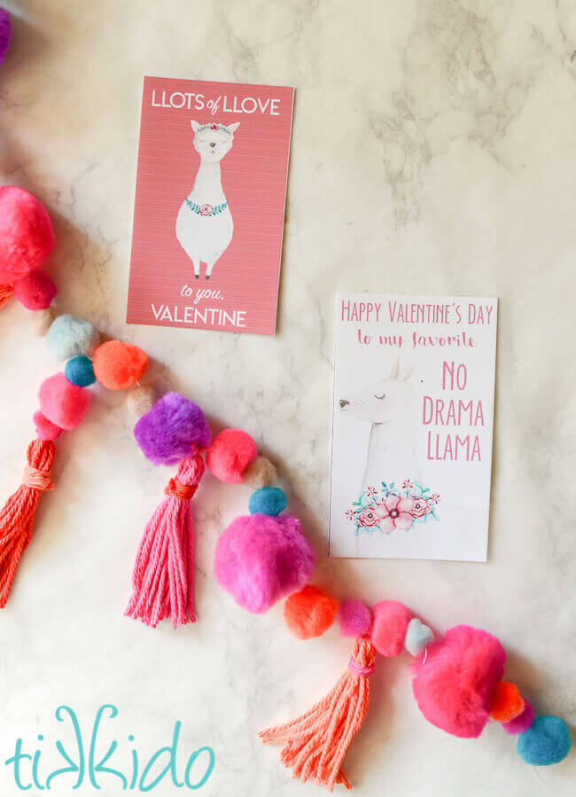 Two free printable llama themed valentines for Valentine's Day next to a colorful pom pom and yarn tassel garland.