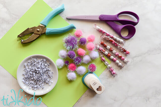 Green cardstock, googly eyes, pom poms, scissors, and sixlet candies on a white marble background.