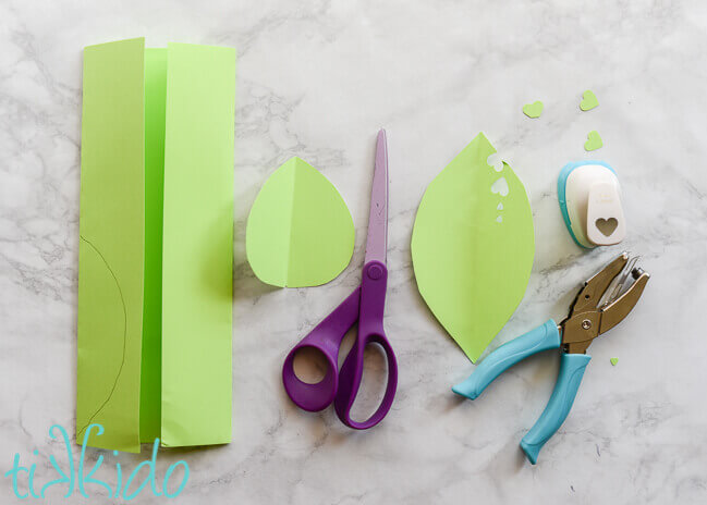 Piece of folded green paper, green paper cut into a leaf shape, scissors, and heart shaped paper punches on white marble background