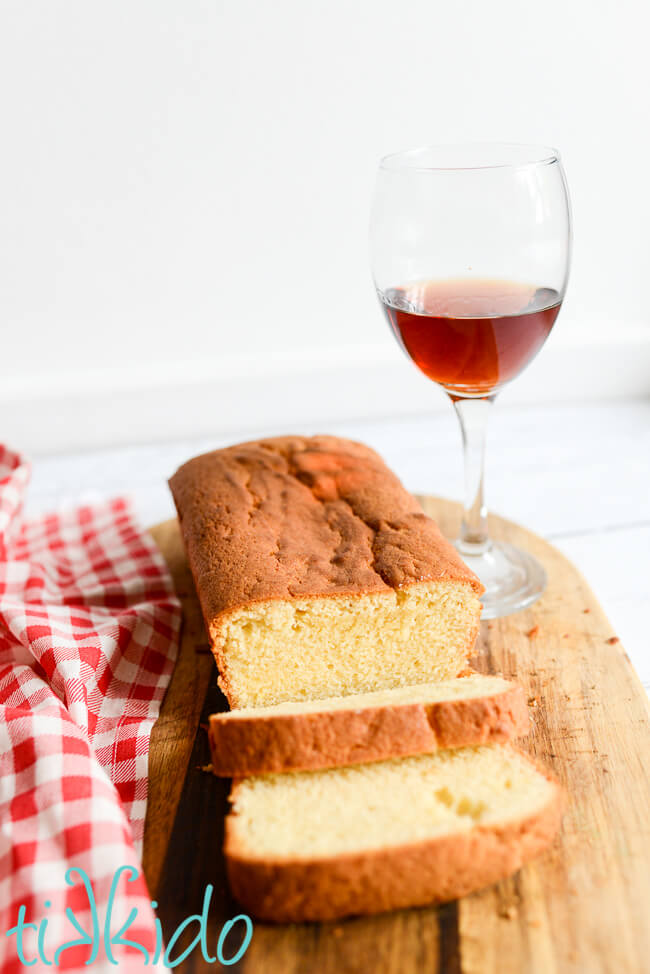 Sliced loaf of Madeira Cake on a wooden cutting board, with a glass of Madeira wine on the right, and a red and white gingham tea towel on the left.