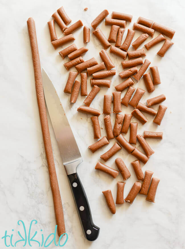 Long roll of homemade tootsie roll, next to sharp knife, next to large grouping of unwrapped tootsie roll candies. 