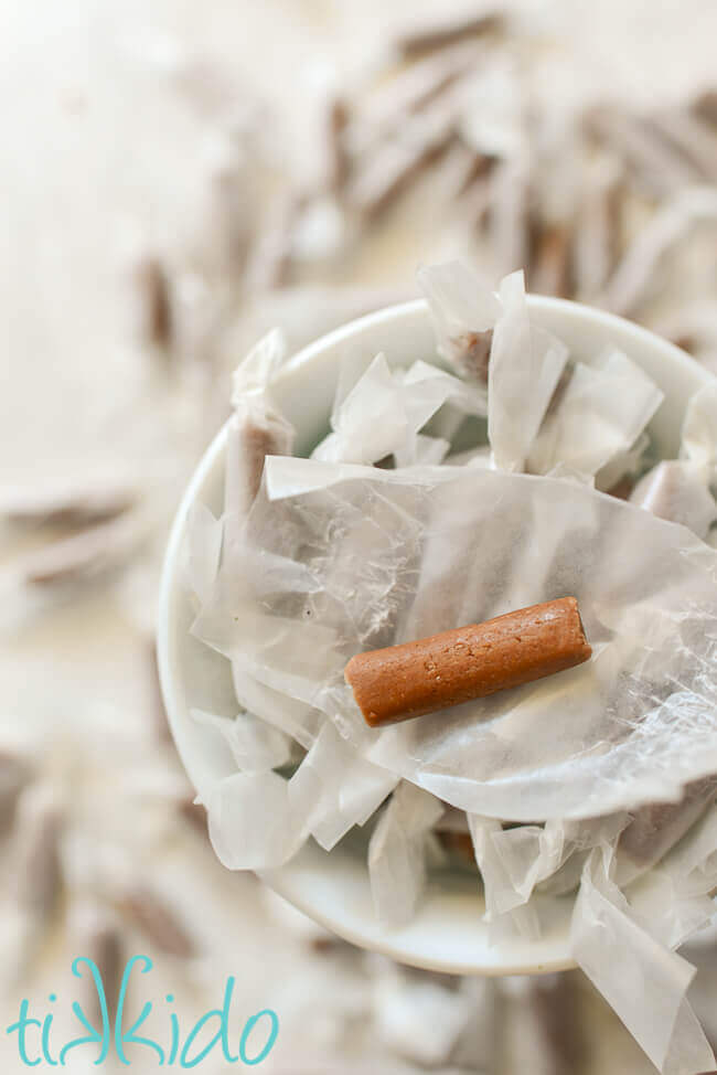 Homemade chocolate malt tootsie rolls wrapped in waxed paper, in a white dish, with one unwrapped tootsie roll sitting on top of the pile.