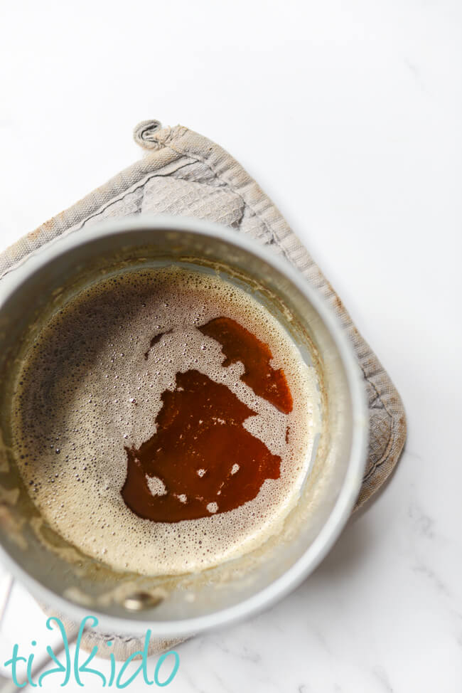 Browned butter for making Browned Butter Frosting in a small saucepan sitting on a grey potholder.