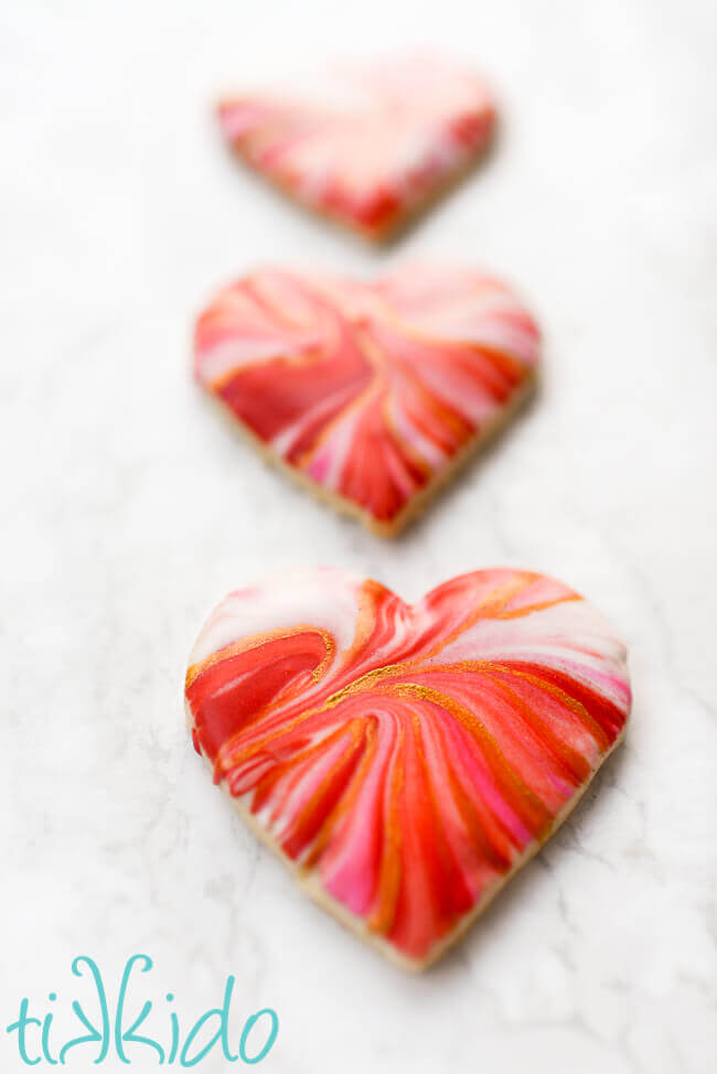 Three  heart shaped sugar cookies dipped in marbled royal icing in reds and pinks and accented with gold edible paint for Valentine's day.