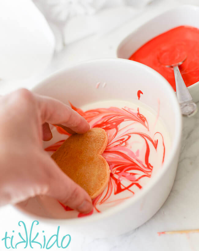 Sugar cookie being dipped in royal icing to create marbled royal icing heart cookies for Valentine's day.