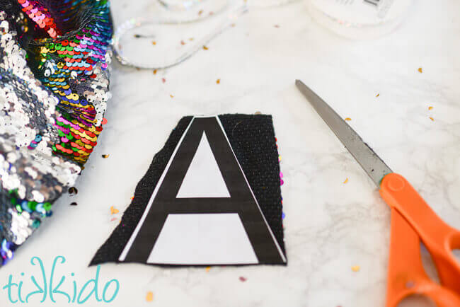 Printed monogram on paper glued to the back side of sparkling mermaid sequin fabric on a white surface.