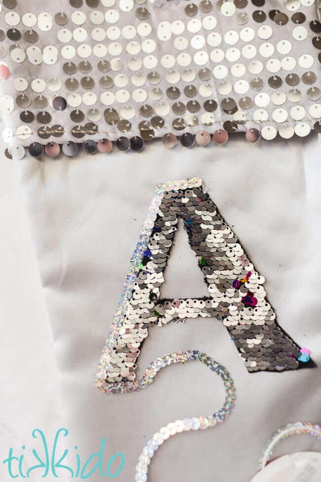 Mermaid sequin fabric monogram on a silver stocking being outlined by silver sequin cord.
