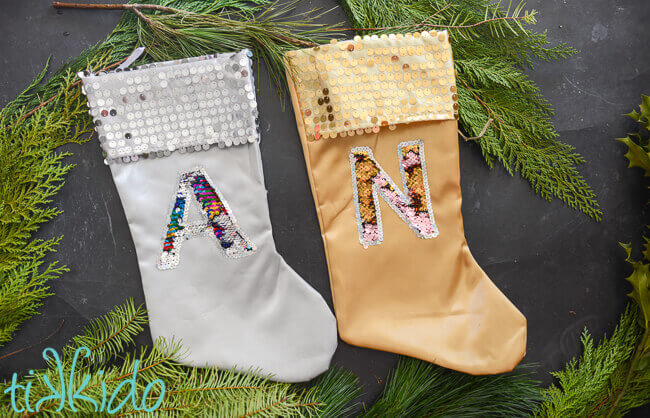 Christmas stockings with mermaid sequin reversible fabric monograms on silver and gold stockings on a black background surrounded by fresh greenery.