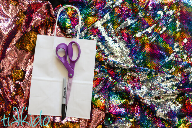 pink and gold and silver and rainbow mermaid sequin fabric and plain white paper gift bag with a pair of scissors and a marker on top.