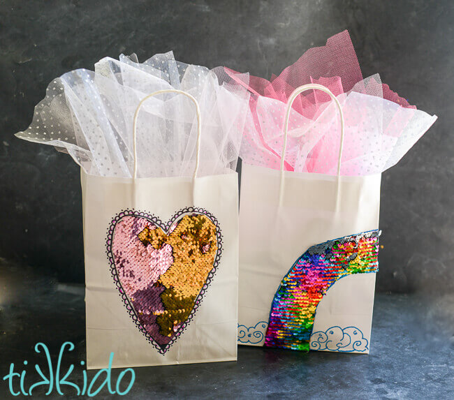 Two white gift bags embellished with sequin mermaid fabric in heart and rainbow shapes.
