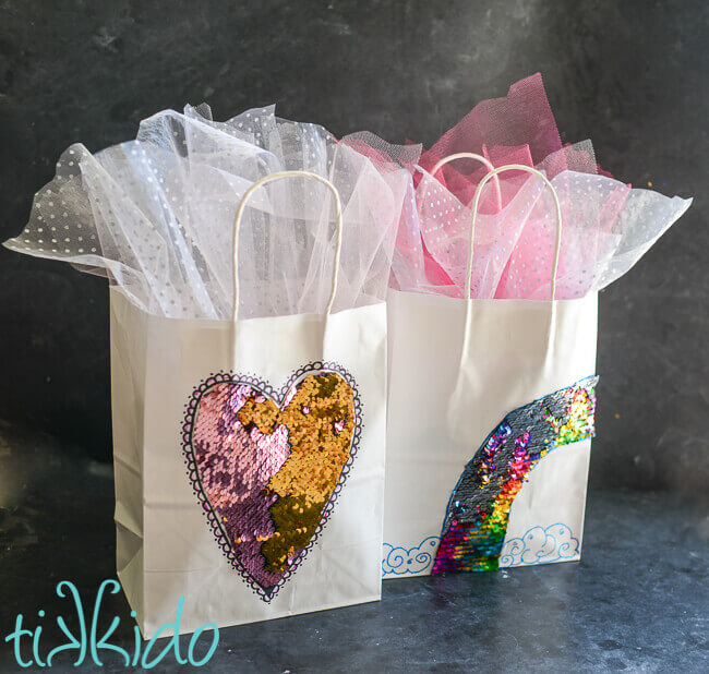 Two white gift bags embellished with sequin mermaid fabric in heart and rainbow shapes.