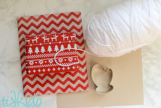 White yarn, cardstock, mitten cookie cutter, and two presents wrapped in red and white nordic sweater print wrapping paper.