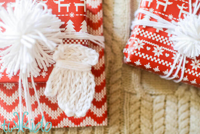 Gift tag that looks like a white knit mitten on a package wrapped with red and white nordic print wrapping paper.