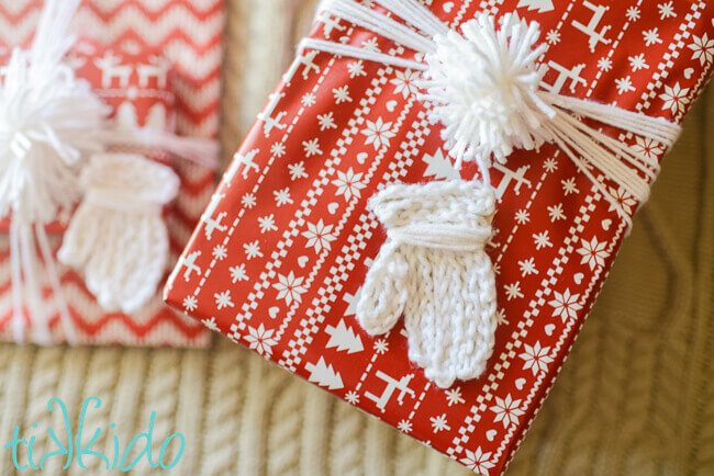 Gift tag that looks like a white knit mitten on a package wrapped with red and white nordic print wrapping paper.