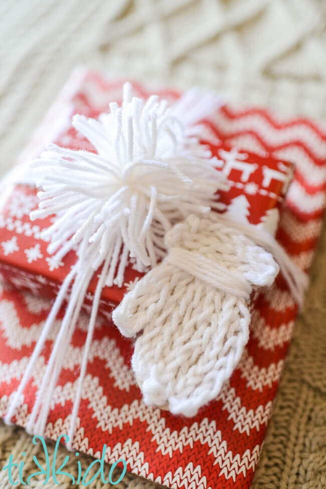 Gift tag that looks like a white knit mitten on a package wrapped with red and white nordic print wrapping paper.