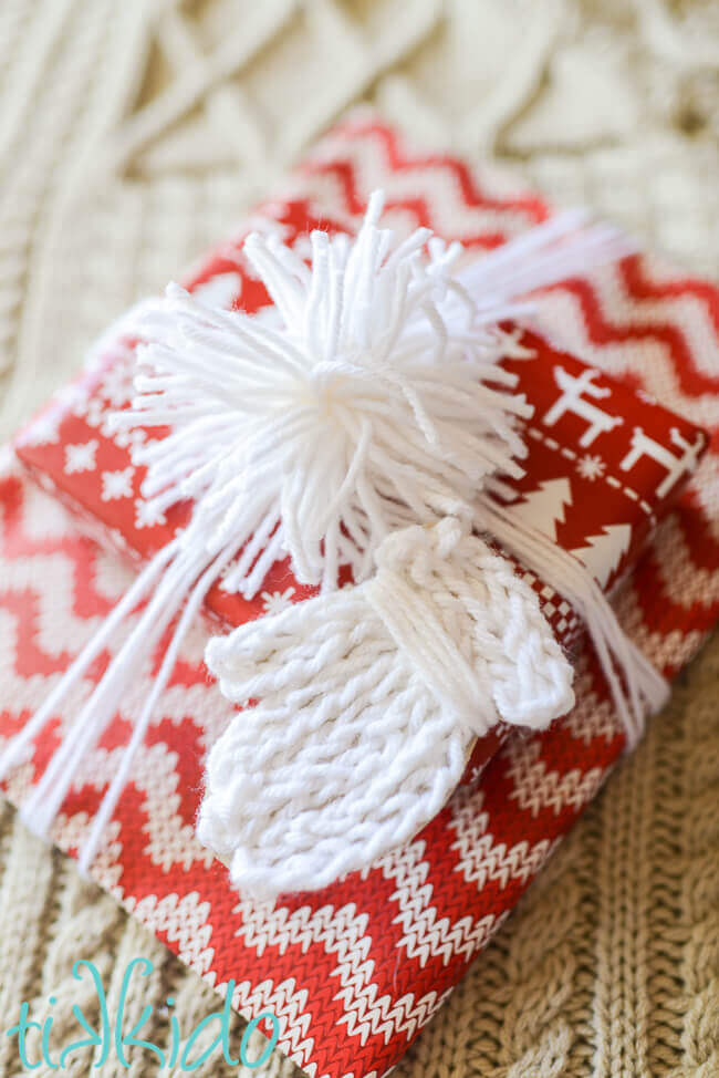 Gift tag that looks like a white knit mitten on a package wrapped with red and white nordic print wrapping paper.