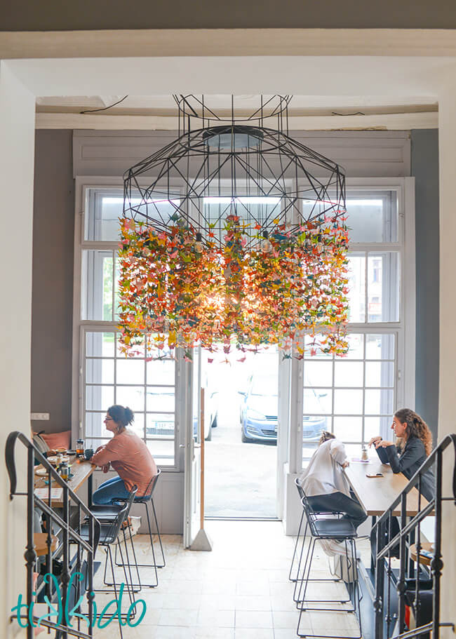 Interior view of SORRY - PEČEME JINAK cafe in Brno, Czech Republic, looking from the cafe toward the front door, featuring a modern chandelier with hundreds of colorful origami cranes.