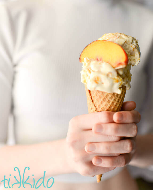Person holding an ice cream cone full of no churn peach ice cream, topped with a slice of peach.