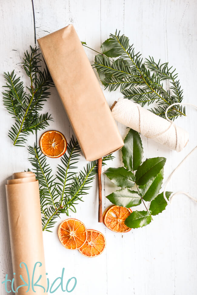 Materials for making a Dried Orange Slice Gift Topper on a white wooden surface.  materials include dried orange slices, cinnamon sticks, string, brown kraft paper, sprigs of holly, and evergreen branches.