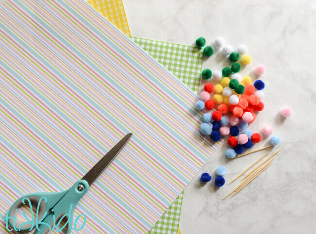 colorful miniature pom poms, green and yellow gingham and striped scrapbook paper, toothpicks, and a pair of aqua scissors on a white marble table surface.