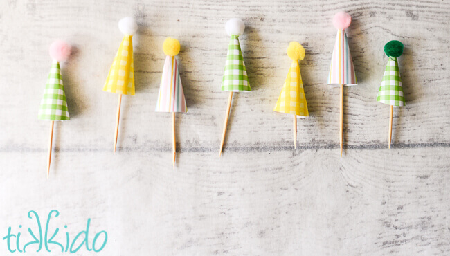 A line of seven miniature paper party hat cupcake toppers on a whitewashed wooden surface.